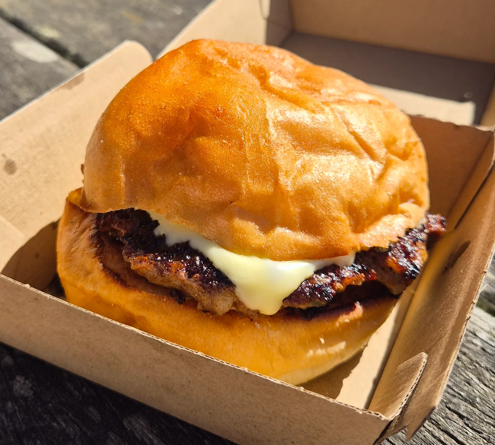 Golden-brown burger with melted cheese in a cardboard takeaway box on a wooden table
